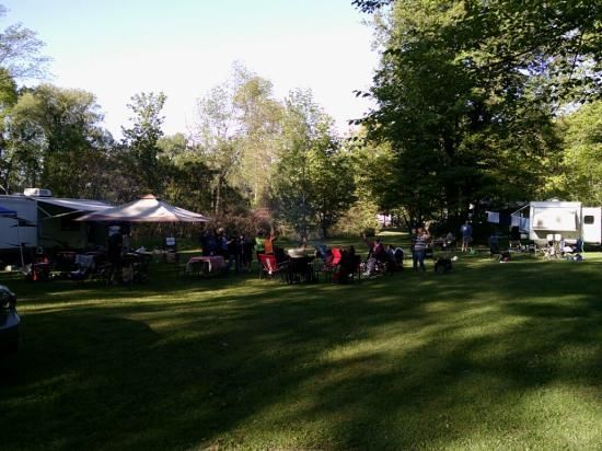 Campground scene. People gather in the grass, near RVs and trees. A tent provides shade. Sunny day.