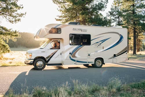 White motorhome parked by a lakeside road, with trees and sunlight in the background.
