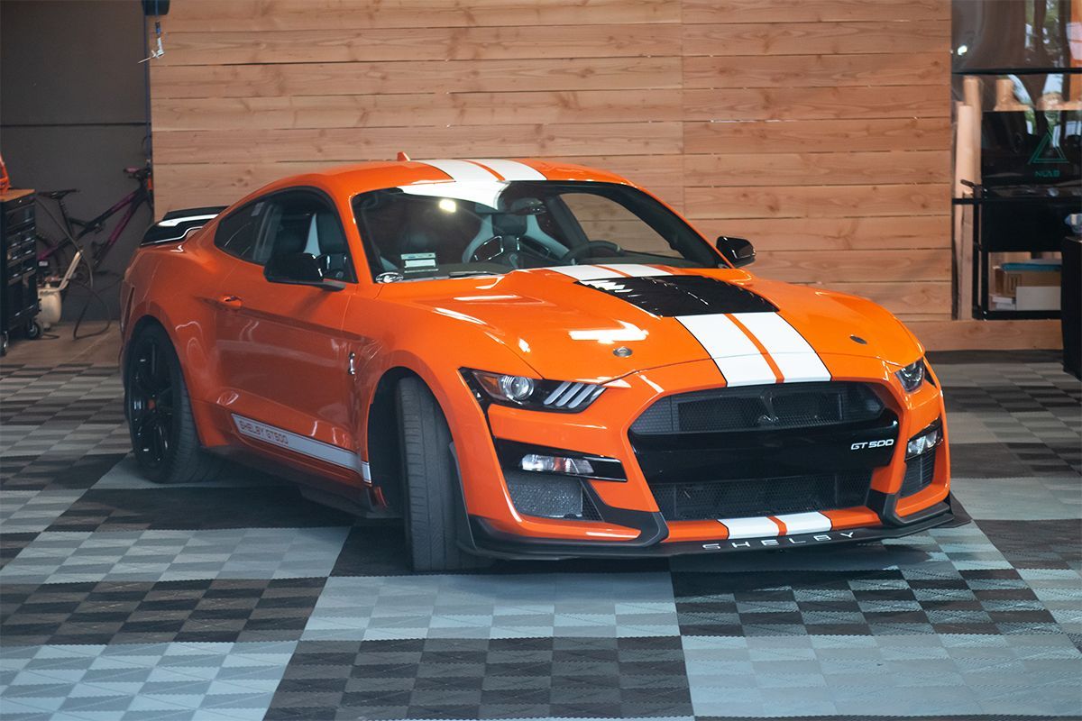 An orange ford mustang is parked on a checkered floor in a garage.