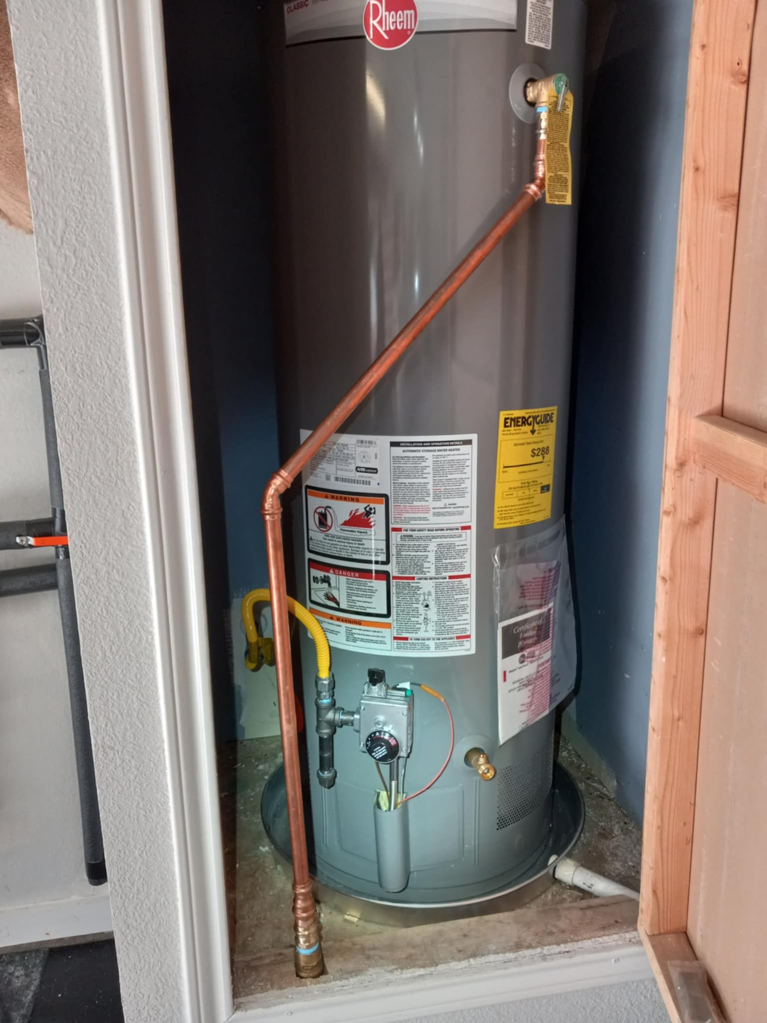 Tall gray water heater with copper piping in a narrow utility closet, viewed through an open doorway