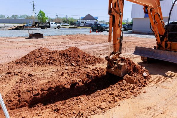 An excavator digging a trench in reddish-brown dirt at a construction site on a sunny day.