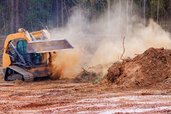 Yellow skid steer mulching wood debris in a clearing, creating a dust cloud.