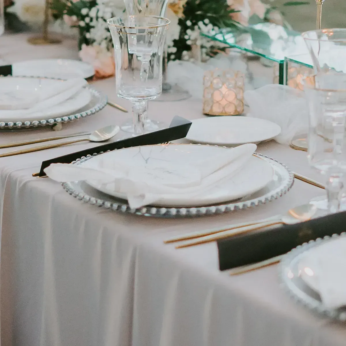 Elegant dining table set with white plates, gold cutlery, folded napkins, and soft floral centerpieces.