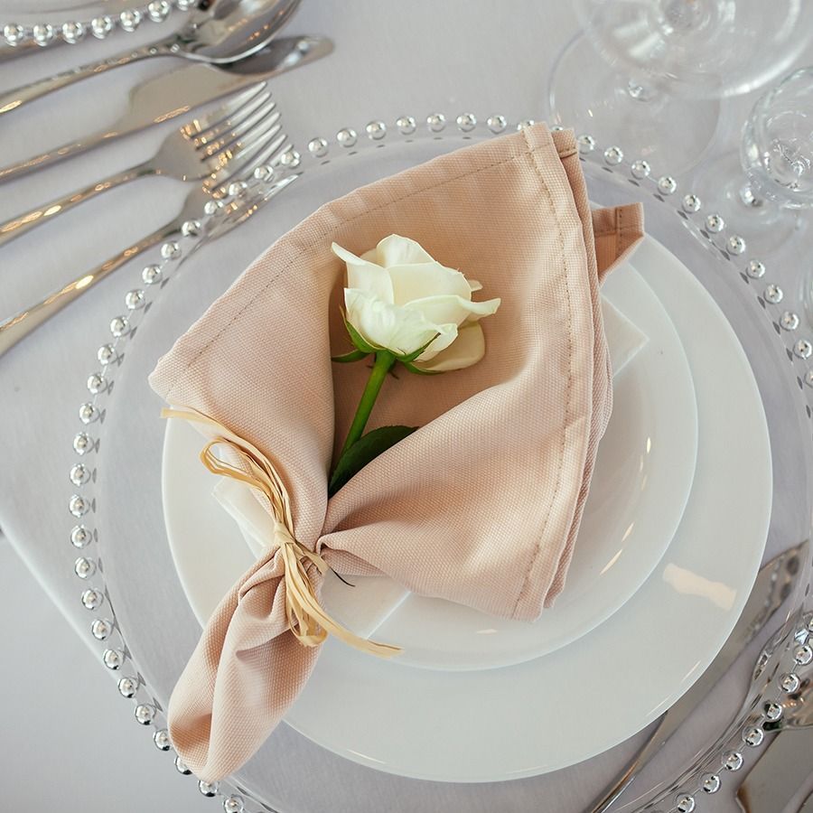 White rose on a folded blush napkin atop a white plate with gold-accented cutlery and pearl beads on a table setting