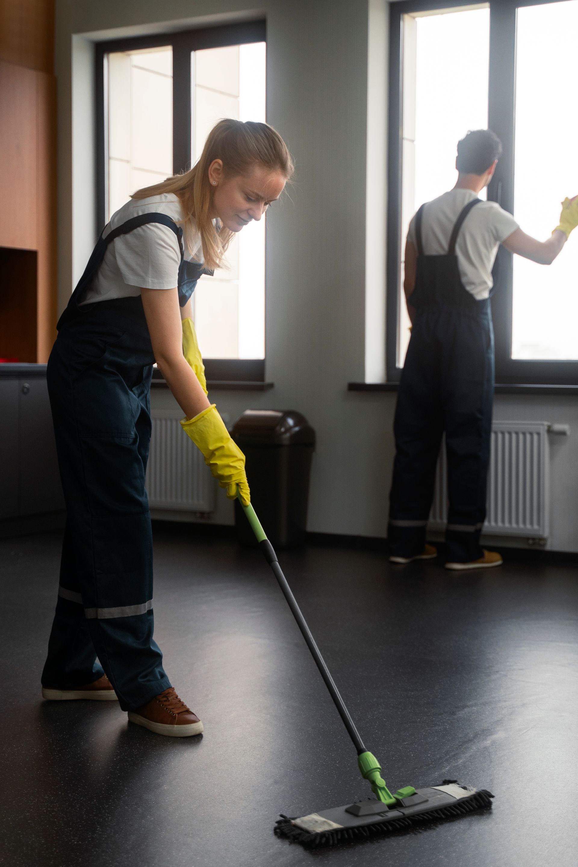 A woman is mopping the floor in a room while a man is cleaning the window.