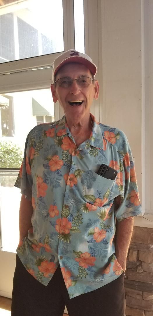 A man wearing a hawaiian shirt and a baseball cap is standing in front of a window.