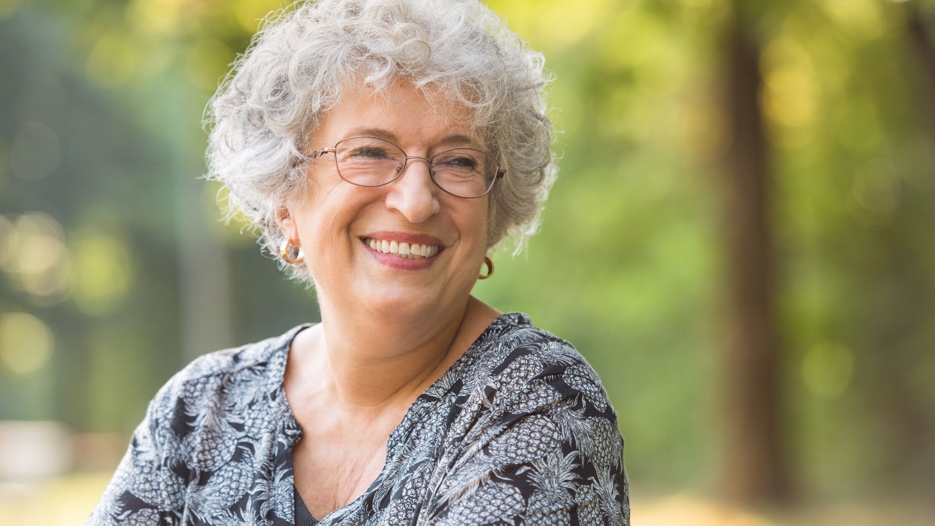 An elderly woman with glasses is smiling in a park.