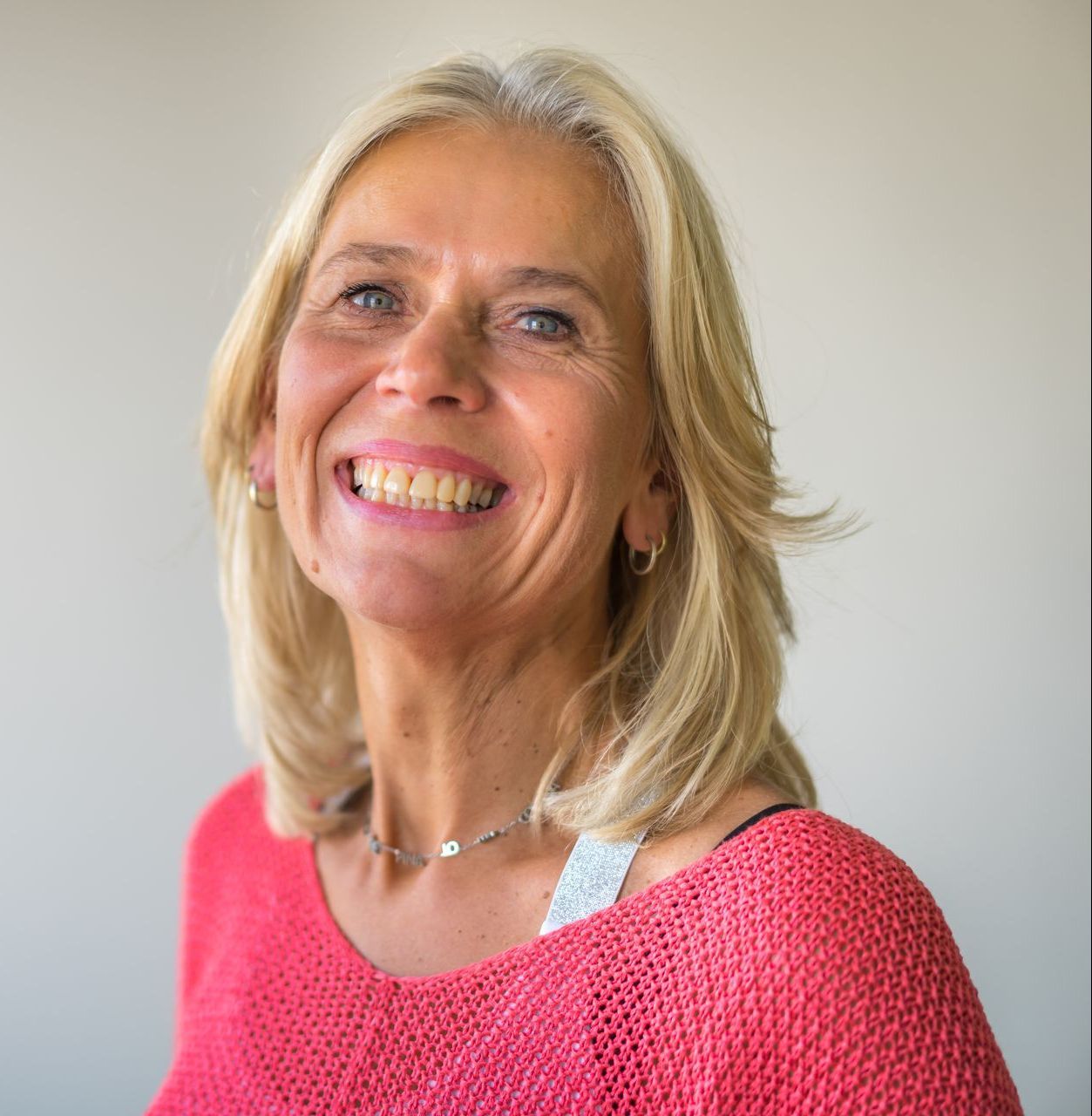 Een blonde vrouw lacht breeduit, gekleed in een koraalkleurige trui, tegen een lichtgrijze achtergrond.