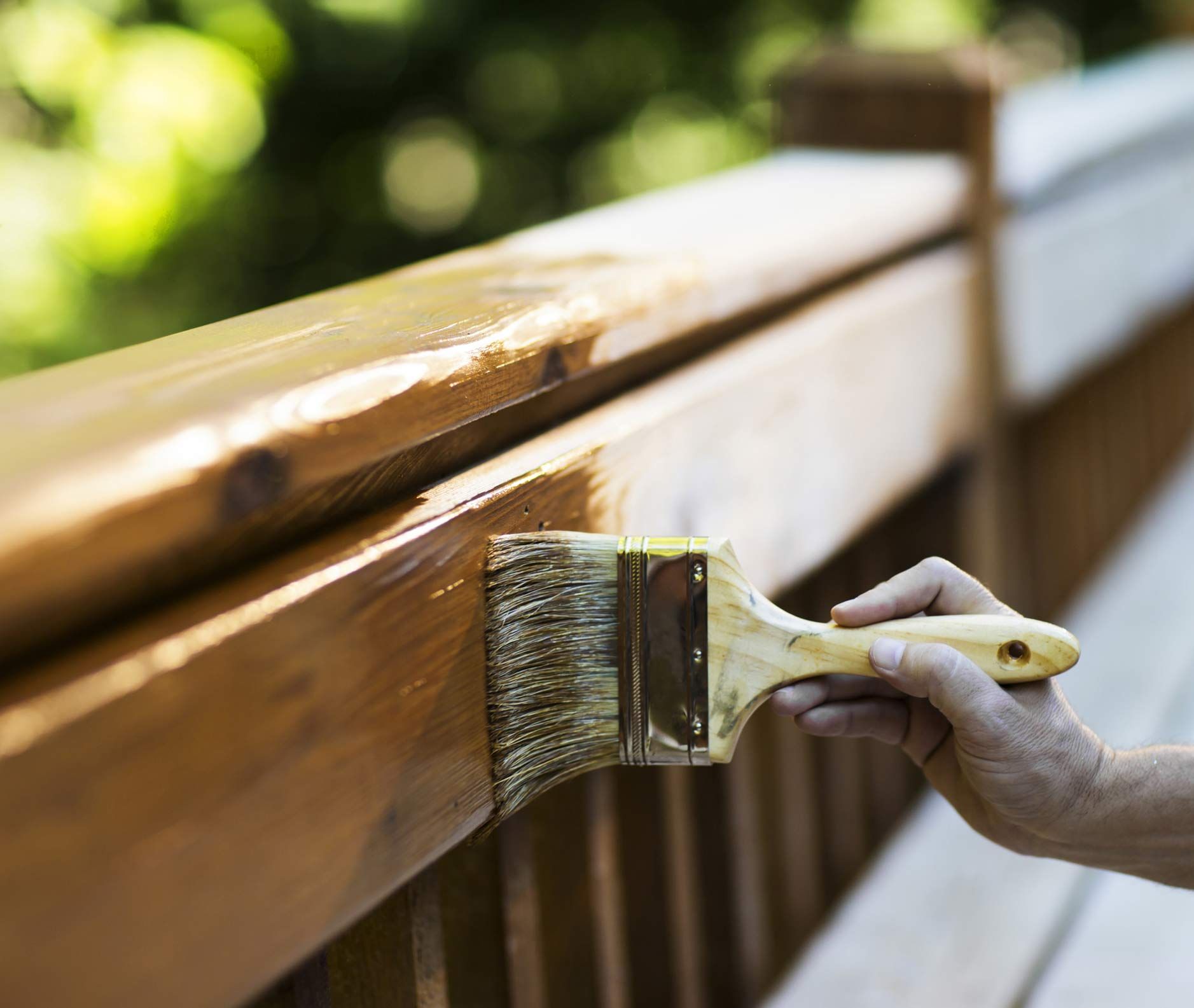Hand applying wood stain to deck railing with a brush in an outdoor setting.