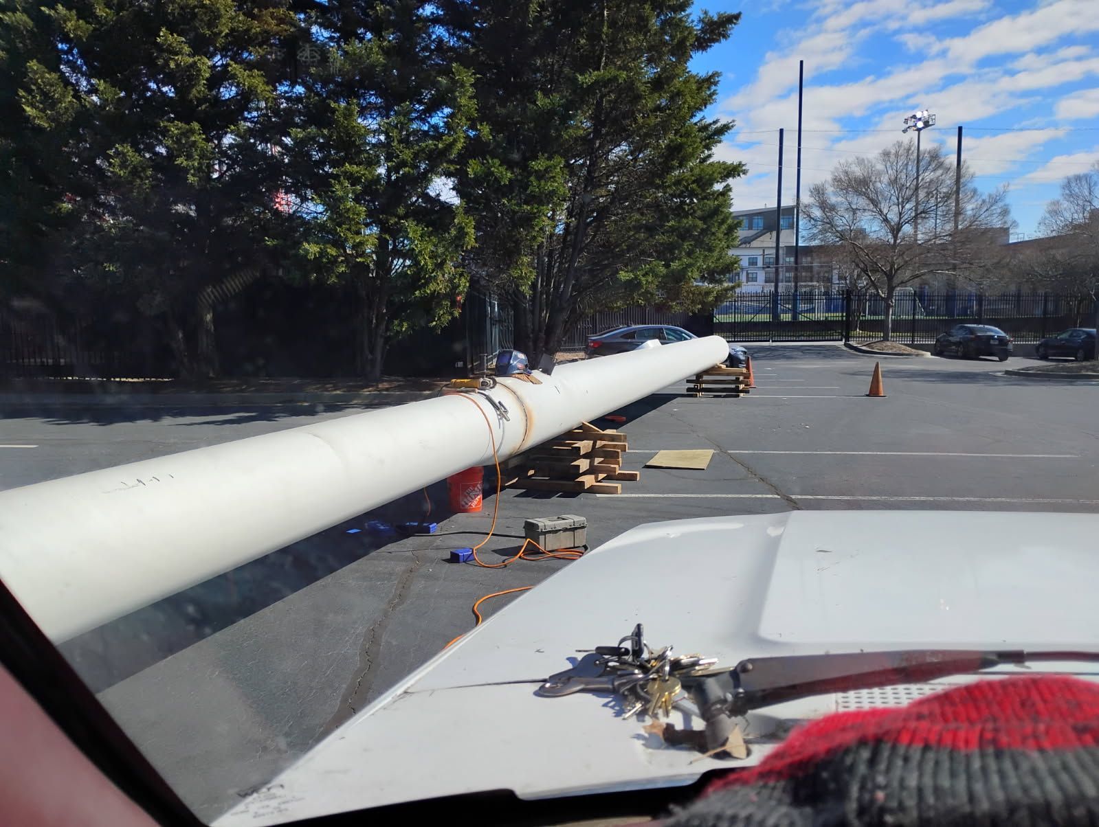 A long white pole extends across a parking lot with traffic cones and trees in the background.