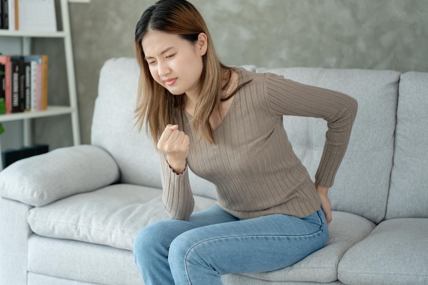 Woman on couch grimaces, clutching lower back.