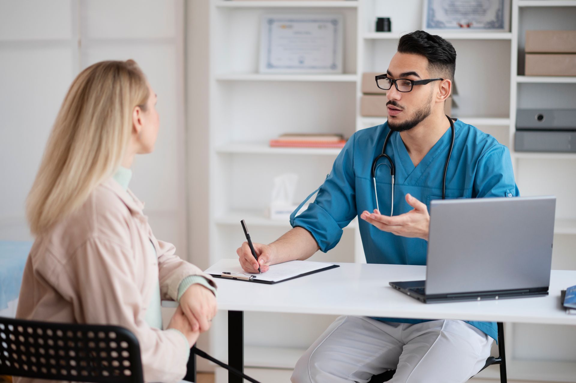 A doctor is talking to a patient while sitting at a desk with a laptop.