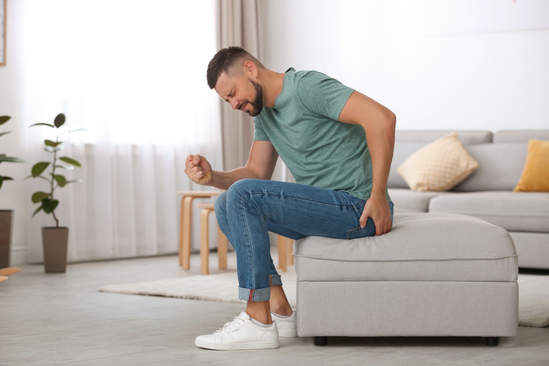 A man is sitting on an ottoman in a living room with his hands on his hips.