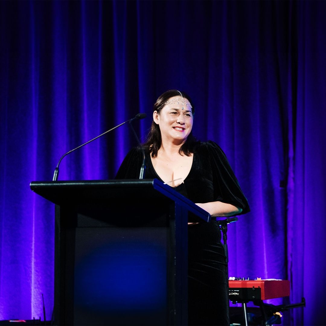 A woman is standing at a podium with a microphone in front of a purple curtain.