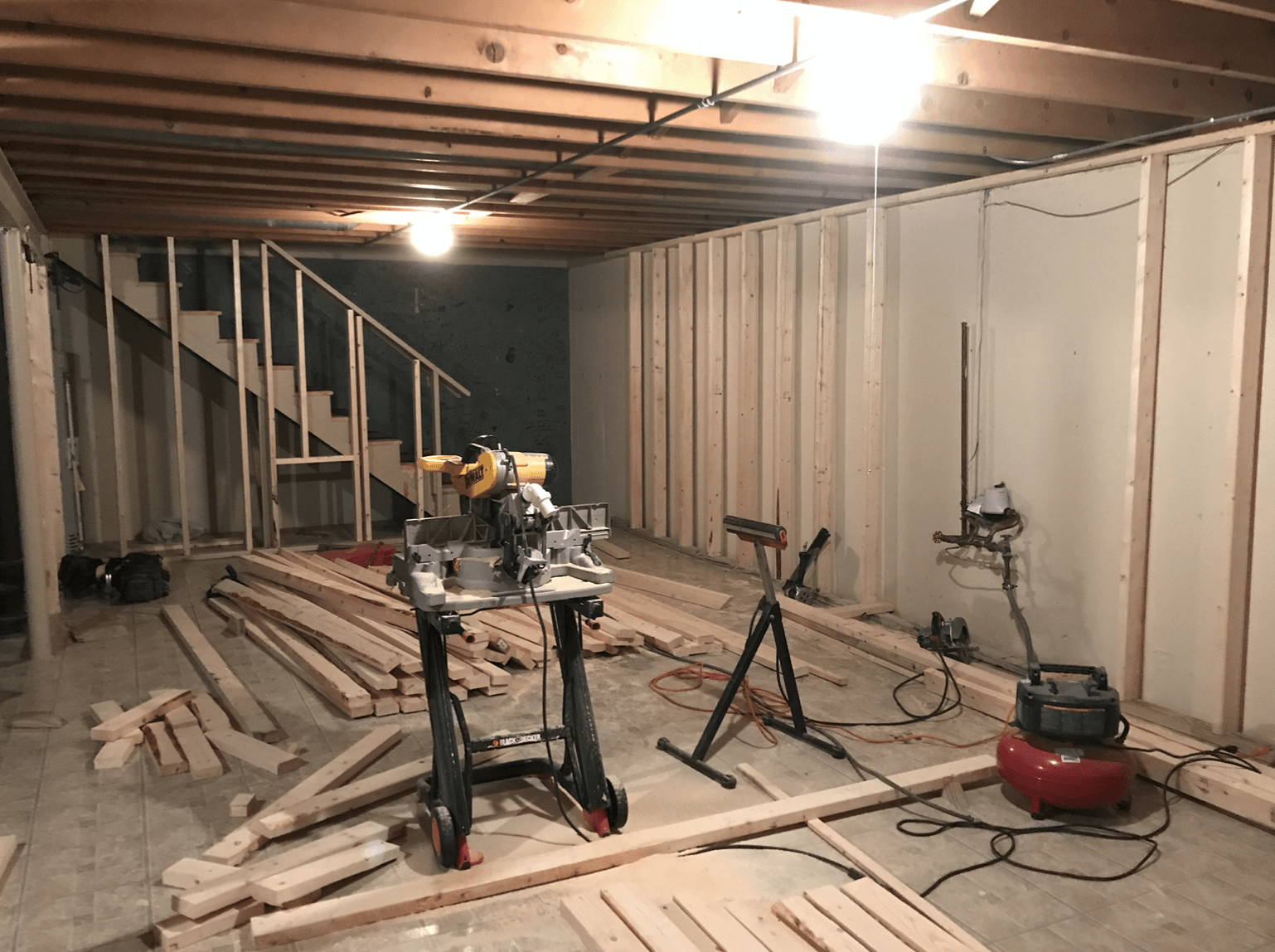 A basement under construction with a table saw , air compressor , and stairs.
