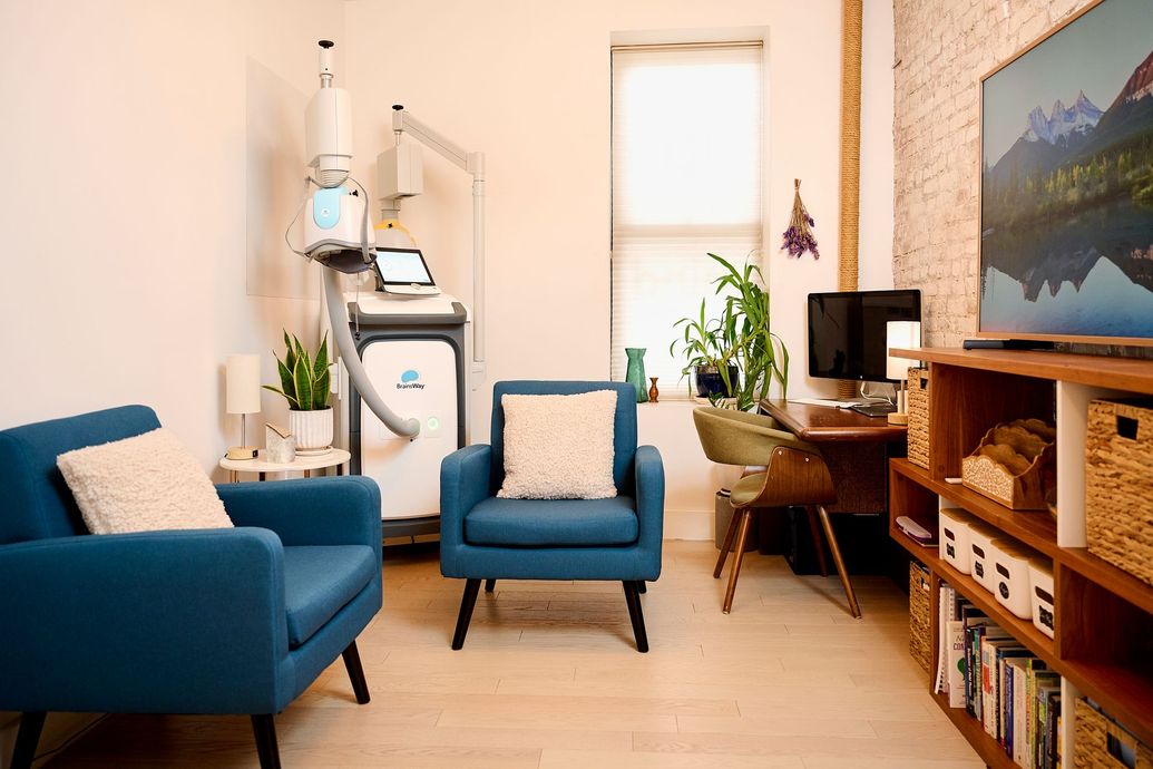A living room with two blue chairs , a desk , a television and a machine.