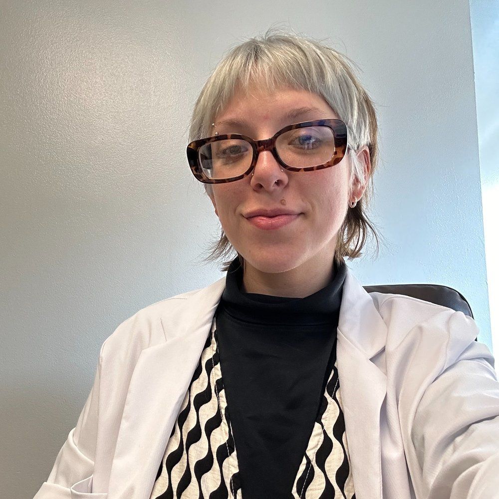 A woman wearing glasses and a white coat is taking a selfie.