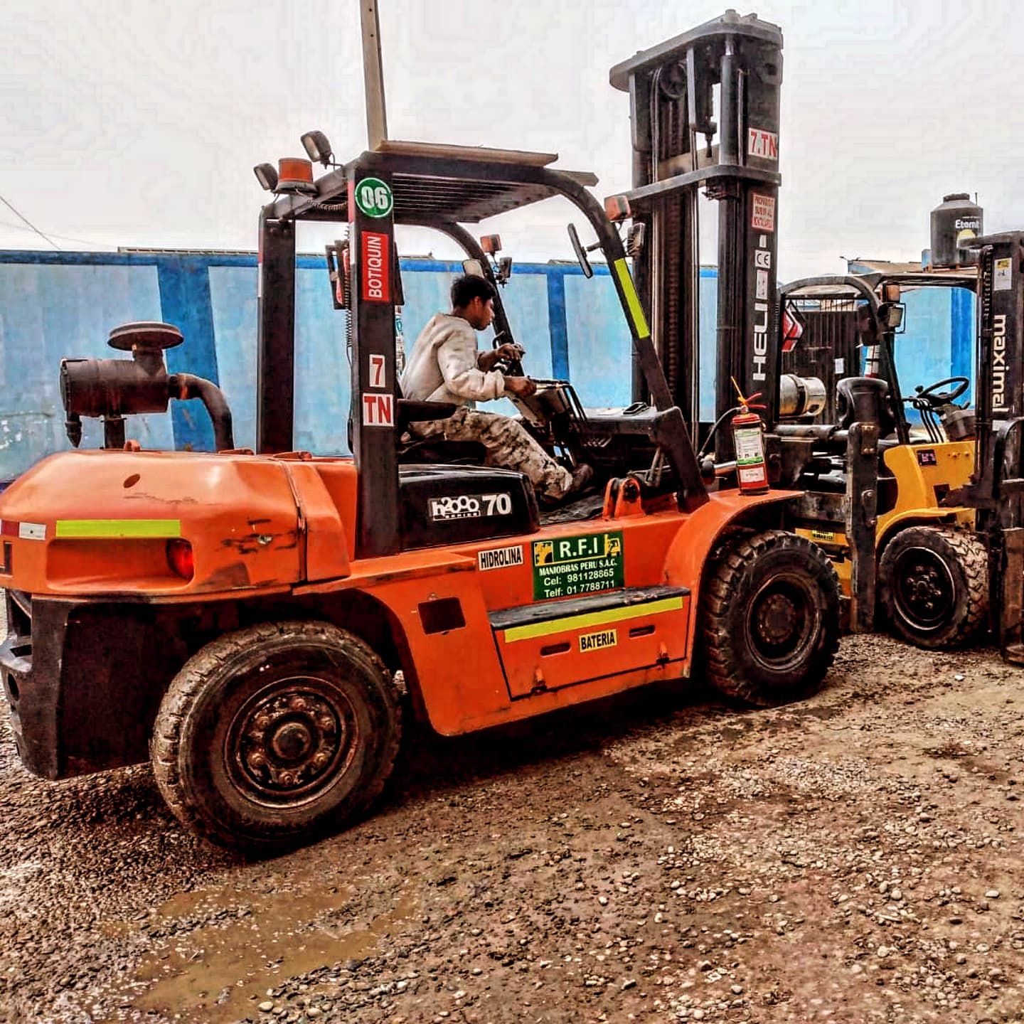 Carretilla elevadora naranja con conductor, otra carretilla al fondo. Suelo embarrado.