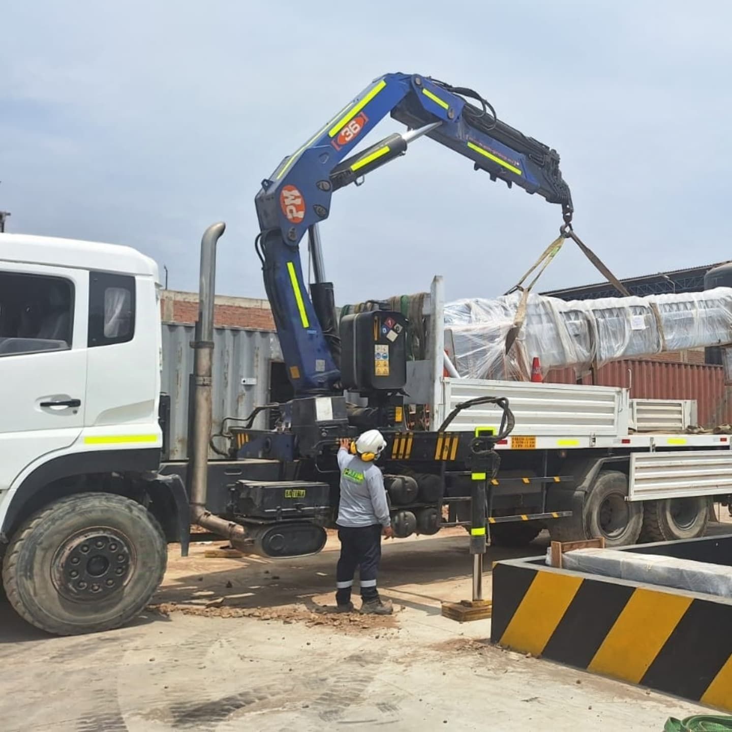 Un trabajador guía una grúa que carga un objeto grande y envuelto en un camión de plataforma en un sitio de construcción.