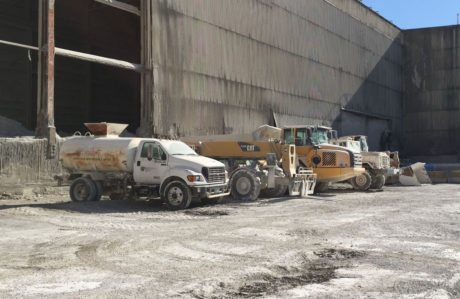 A water truck and construction vehicles parked outside a large industrial building.