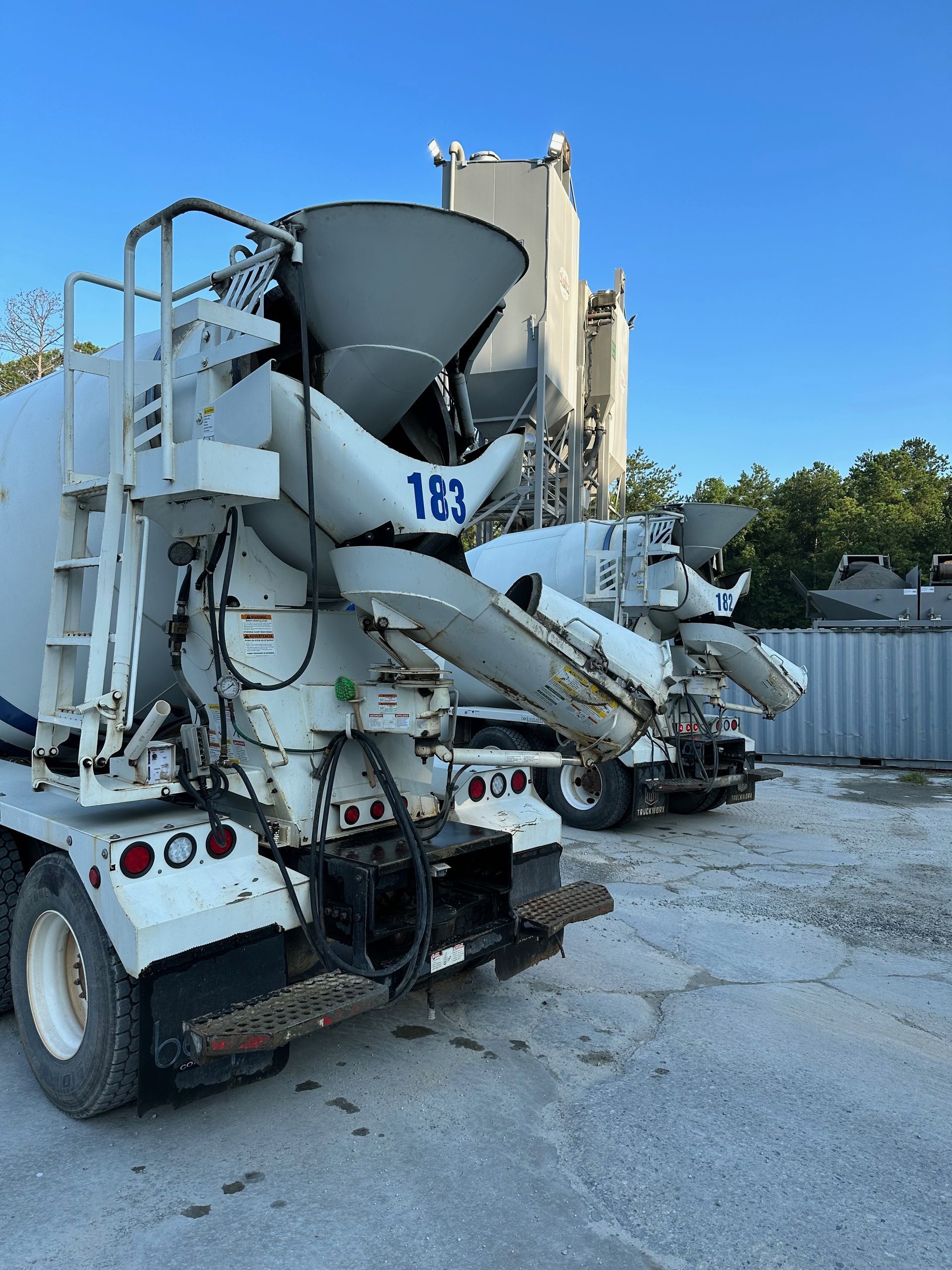Two white cement mixer trucks parked outdoors near a concrete plant, under a blue sky.