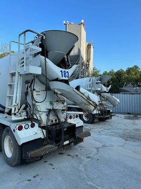 Two white cement mixer trucks parked outdoors near a concrete plant, under a blue sky.