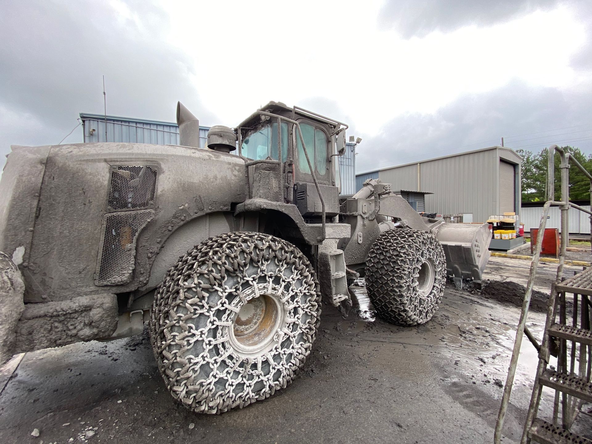 Mud-covered loader with tire chains near a building, cloudy sky.