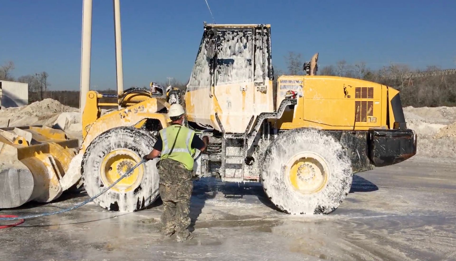 A person in a vest cleaning a large, yellow construction vehicle covered in foam. Outdoor setting.