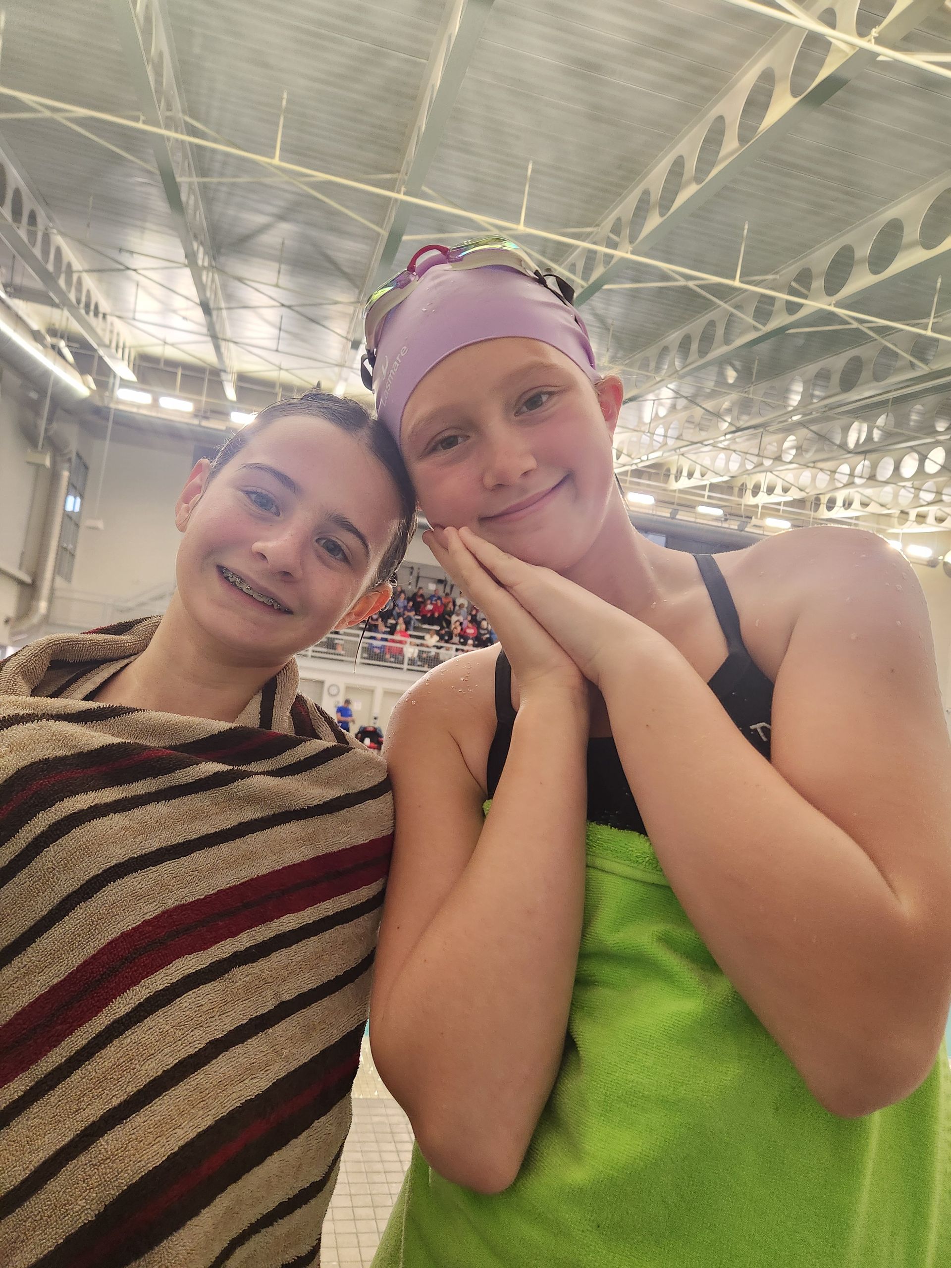 Two smiling swimmers wrapped in towels at an indoor pool. One wears a swim cap and makes a sleepy face.