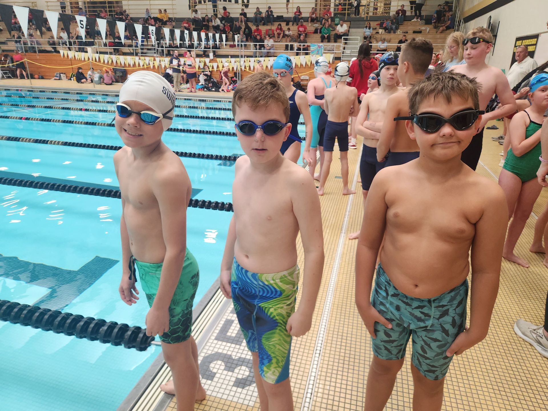 Three young swimmers by a pool, wearing swimsuits and goggles, waiting.