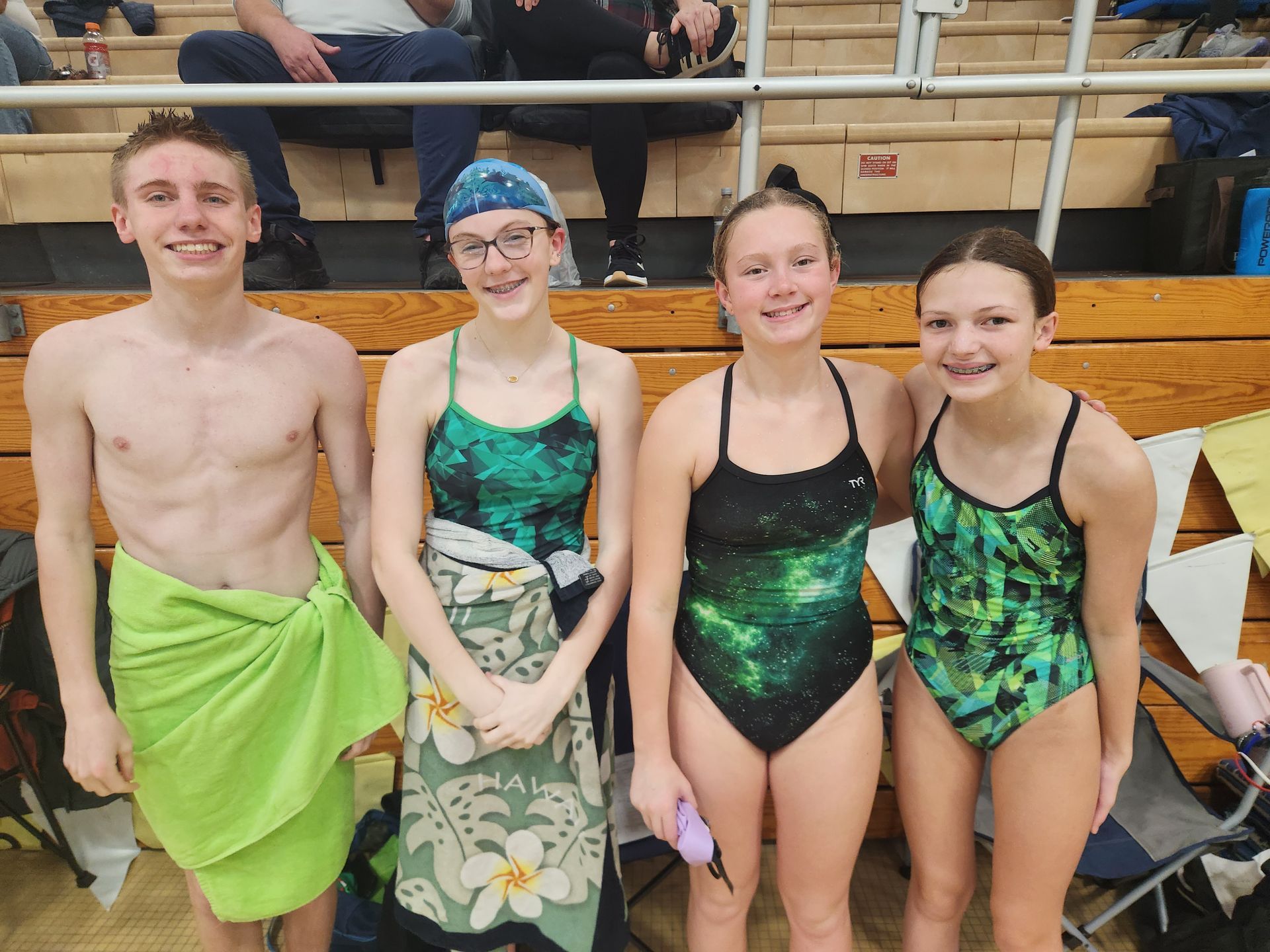 Four swimmers pose after a competition: one with a towel, three in swimsuits, in a pool area.