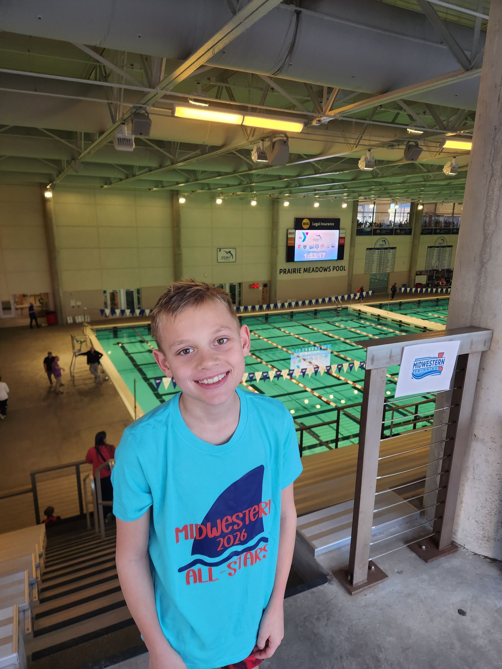 Boy smiling at the camera in front of an indoor swimming pool. He is wearing a blue t-shirt with a shark design.