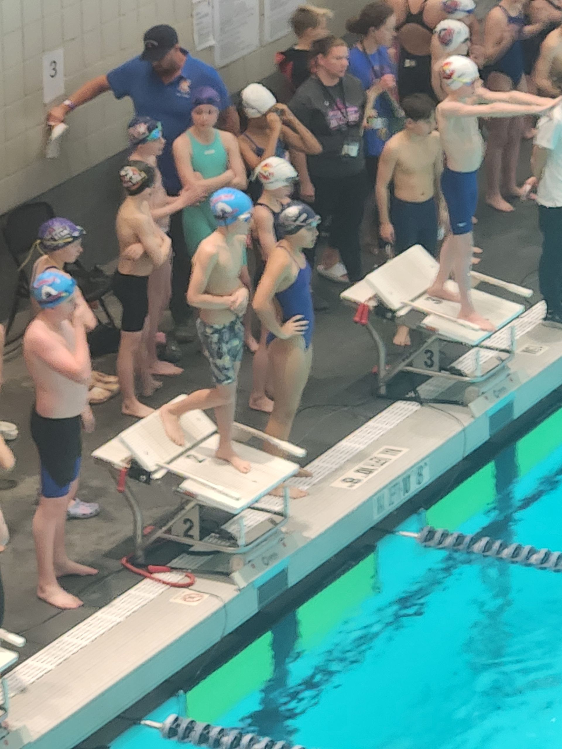 Swimmers on starting blocks at a pool. Some wear swim caps and goggles; spectators watch.