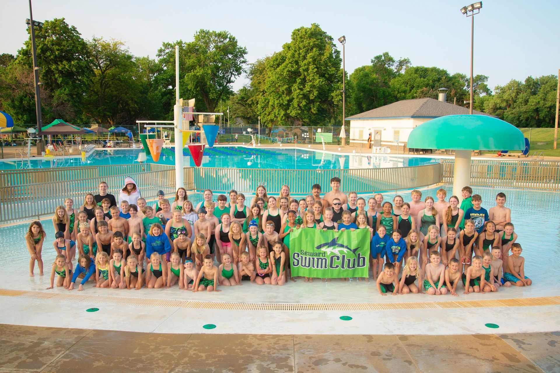Large group of young swimmers in green and blue, posing by a pool with a banner.
