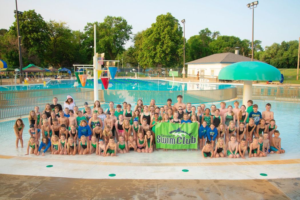 Large group of young swimmers in green and blue, posing by a pool with a banner.