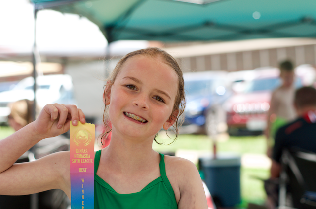Girl smiles, holding a blue, pink, and yellow ribbon at an outdoor event. She wears a green top.