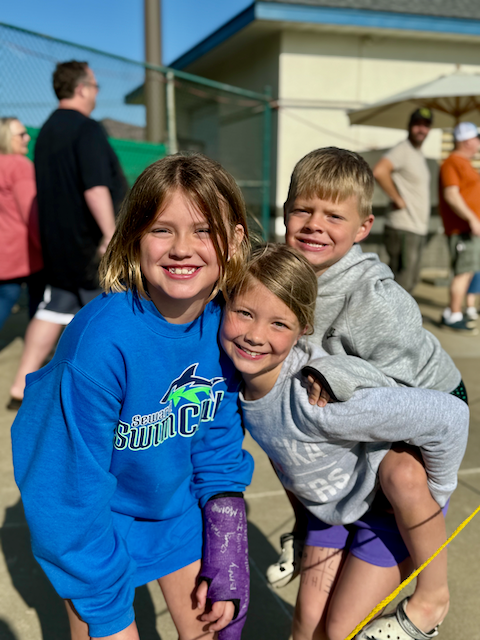 Three smiling children outside: a girl with a cast, and two others giving piggyback rides.