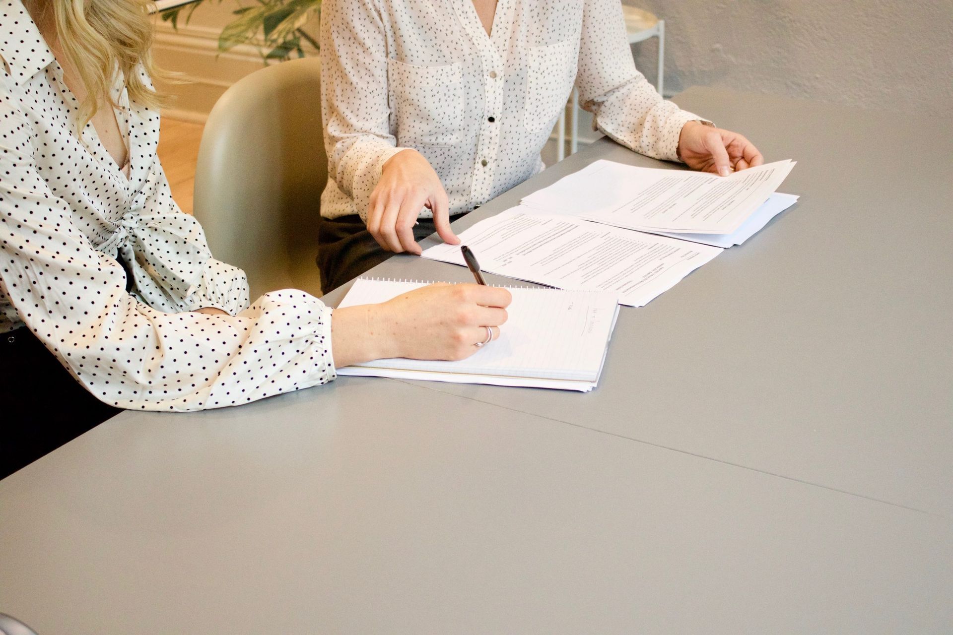 Two women are helping eachother out with documents while seated at a table.
