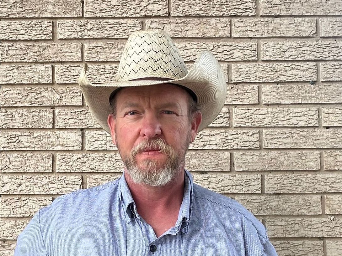 A man with a beard and a hat is standing in front of a brick wall.