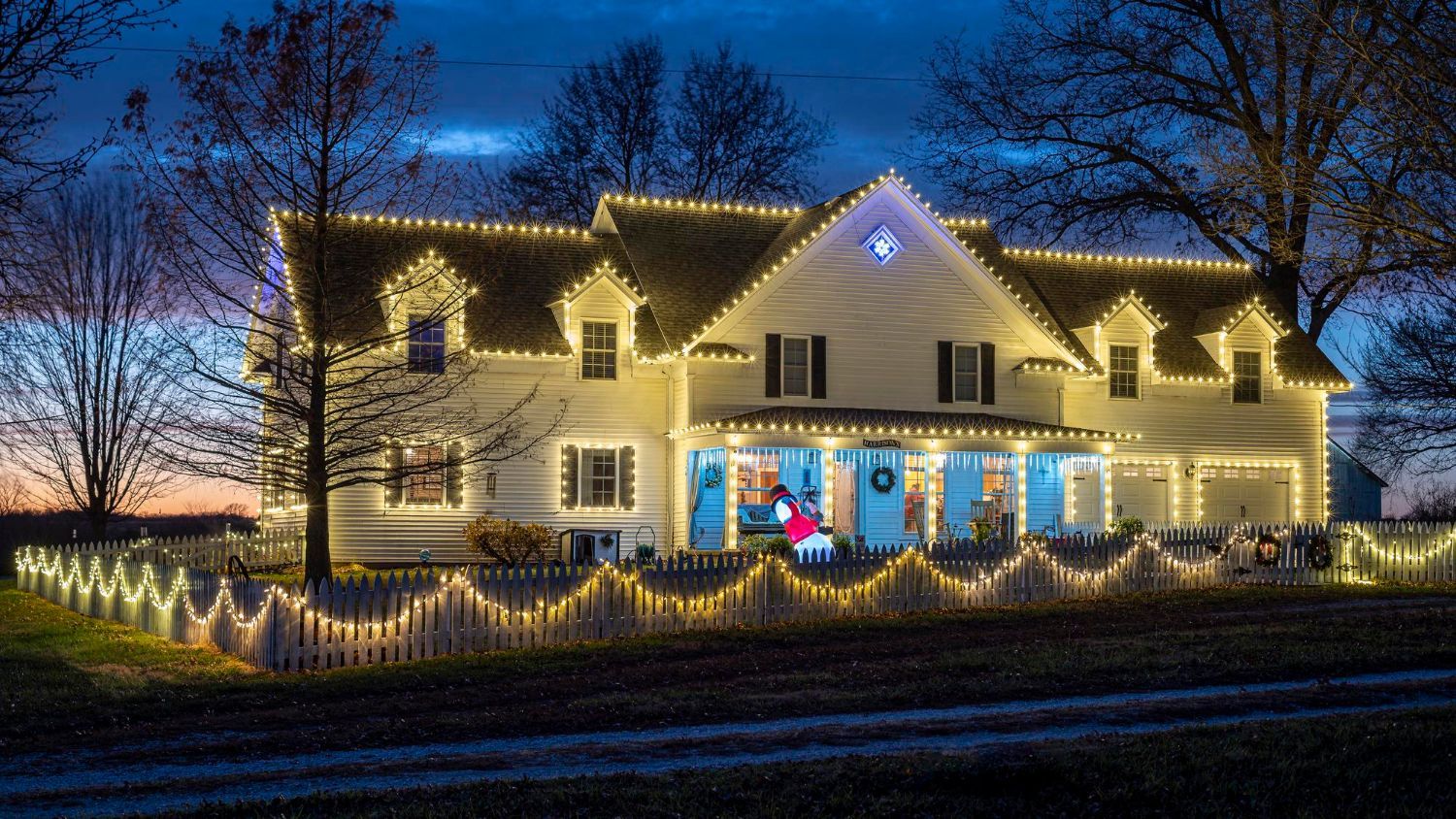 House with white facade, decorated with Christmas lights on the roof, fence, and porch. Evening scene.