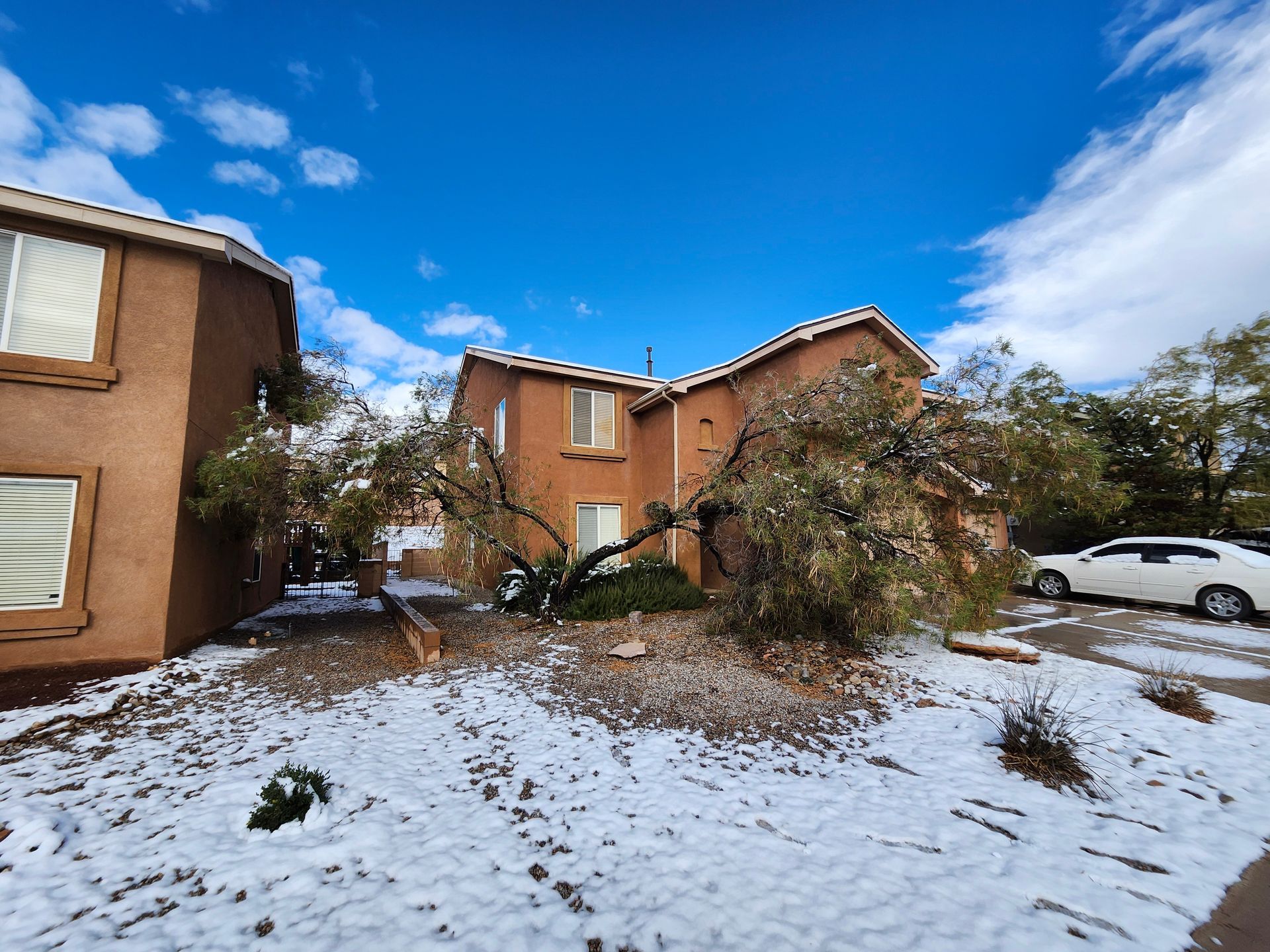 Snow-covered residential neighborhood with brown stucco houses under a blue sky, trees with sparse leaves.