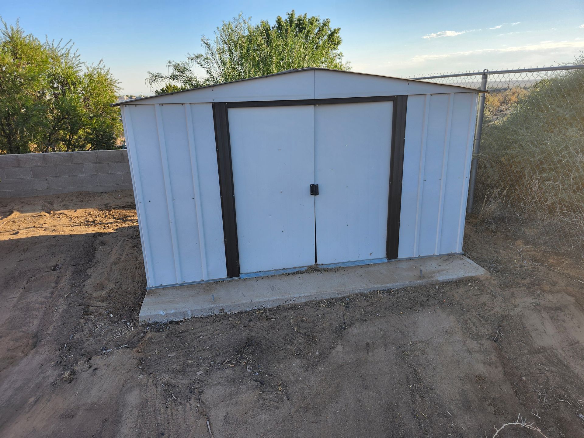 A white metal shed with brown trim sits in the dirt
