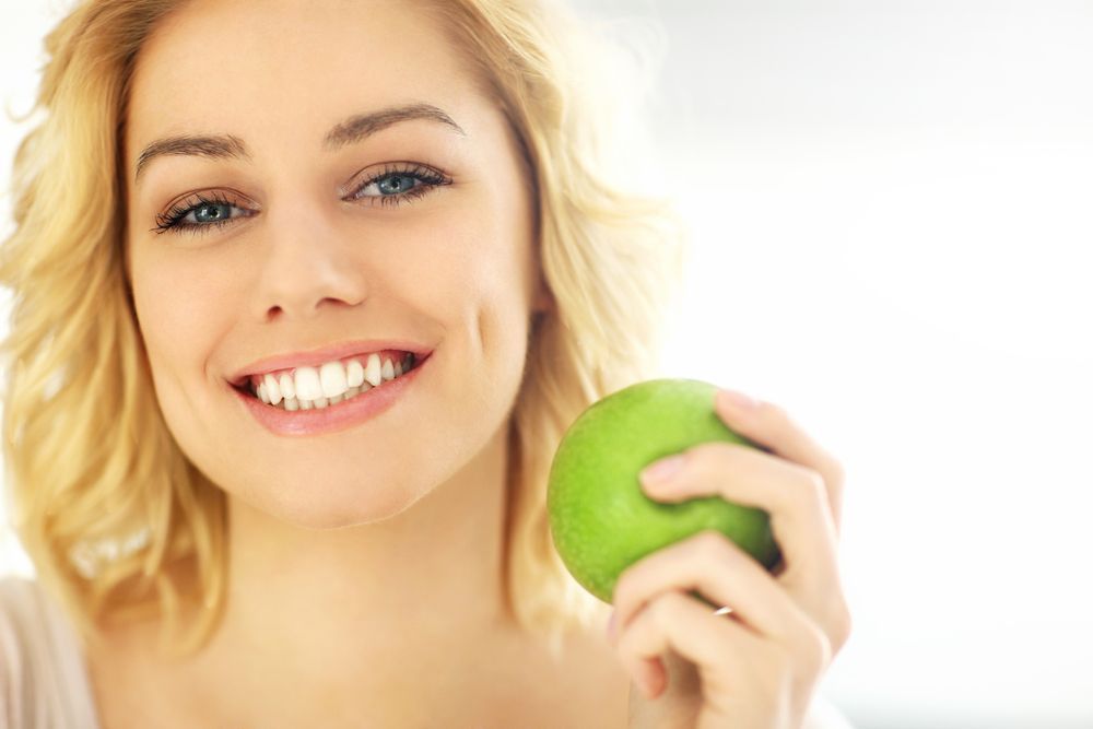 A Woman Is Holding A Green Apple In Her Hand And Smiling — Cairns Precision Dental Group In Woree, QLD