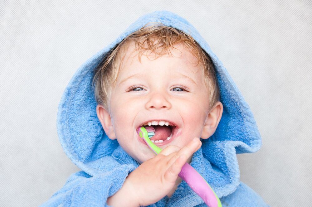 A Young Boy Wearing a Blue Robe is Brushing His Teeth With a Pink Toothbrush — Cairns Precision Dental Group In Woree, QLD