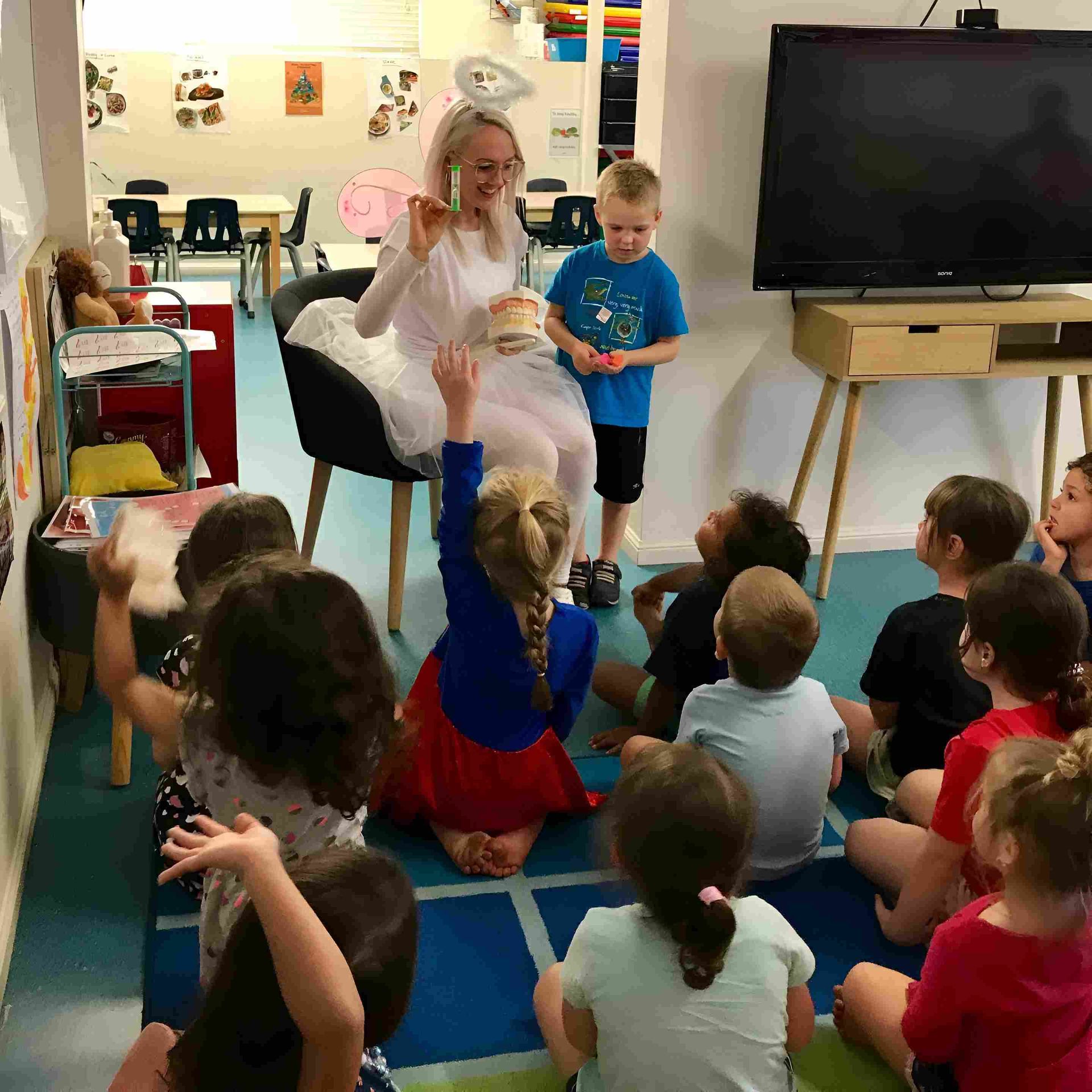 A Woman in a White Dress is Giving a Presentation to a Group of Children — Cairns Precision Dental Group In Woree, QLD