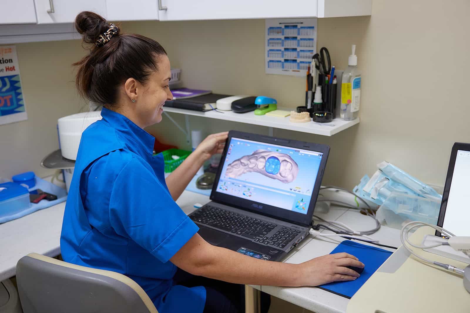 A Woman Is Sitting At A Desk Using A Laptop Computer — Cairns Precision Dental Group In Woree, QLD
