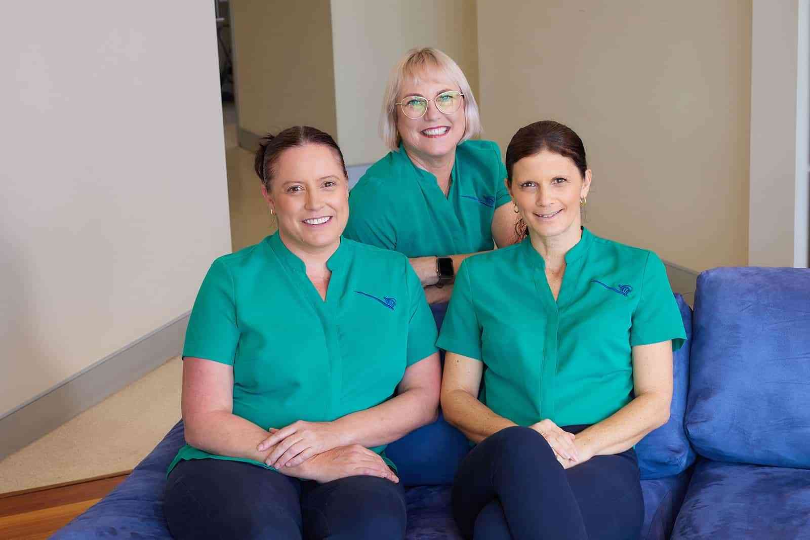 Three Women in Green Scrubs Are Sitting on a Blue Couch — Cairns Precision Dental Group In Woree, QLD
