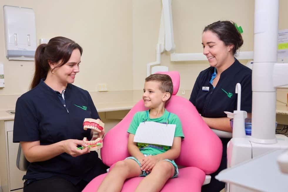 A Young Boy is Sitting in a Pink Dental Chair With Two Female Dentists — Cairns Precision Dental Group In Woree, QLD