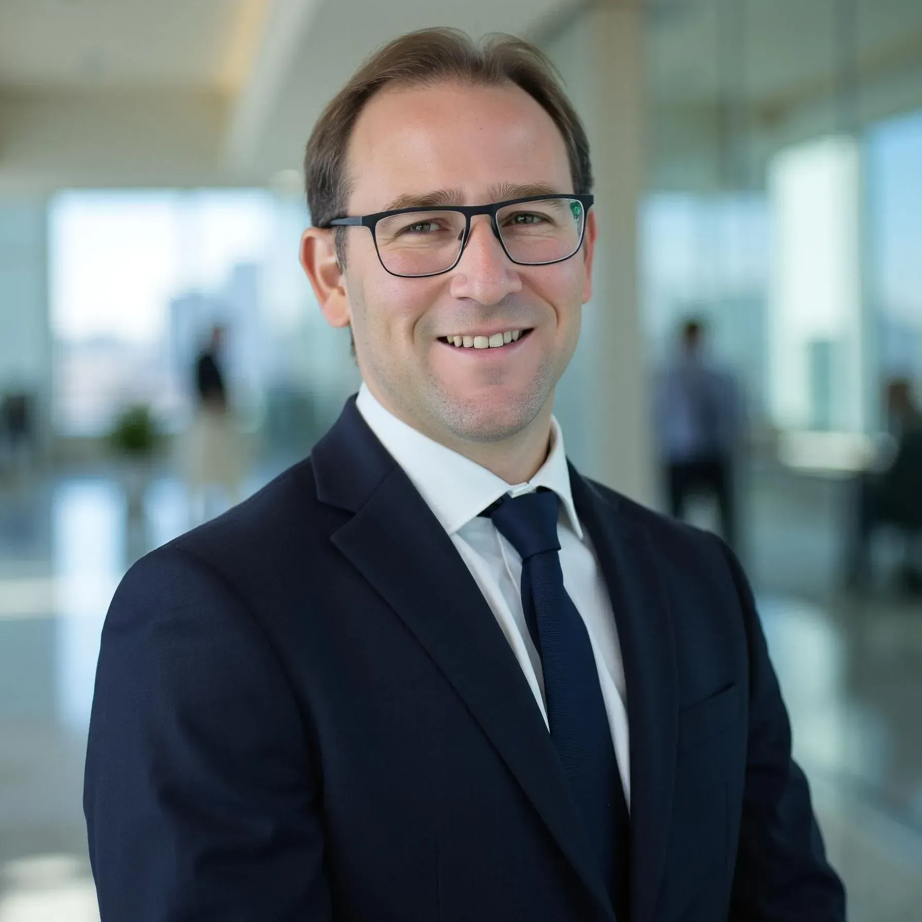 Man in navy suit, white shirt, smiling against a gray backdrop.