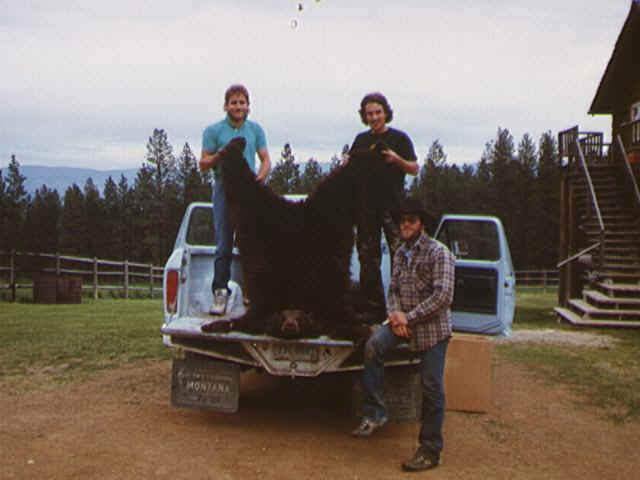 two men standing next to a large black bear in the back of a truck